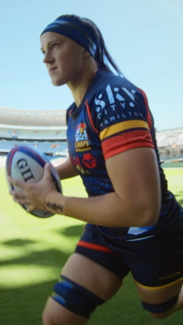 Female rugby player in uniform runs with the ball in an outdoor stadium, blue sky background, athletic sportswear, determined expression.