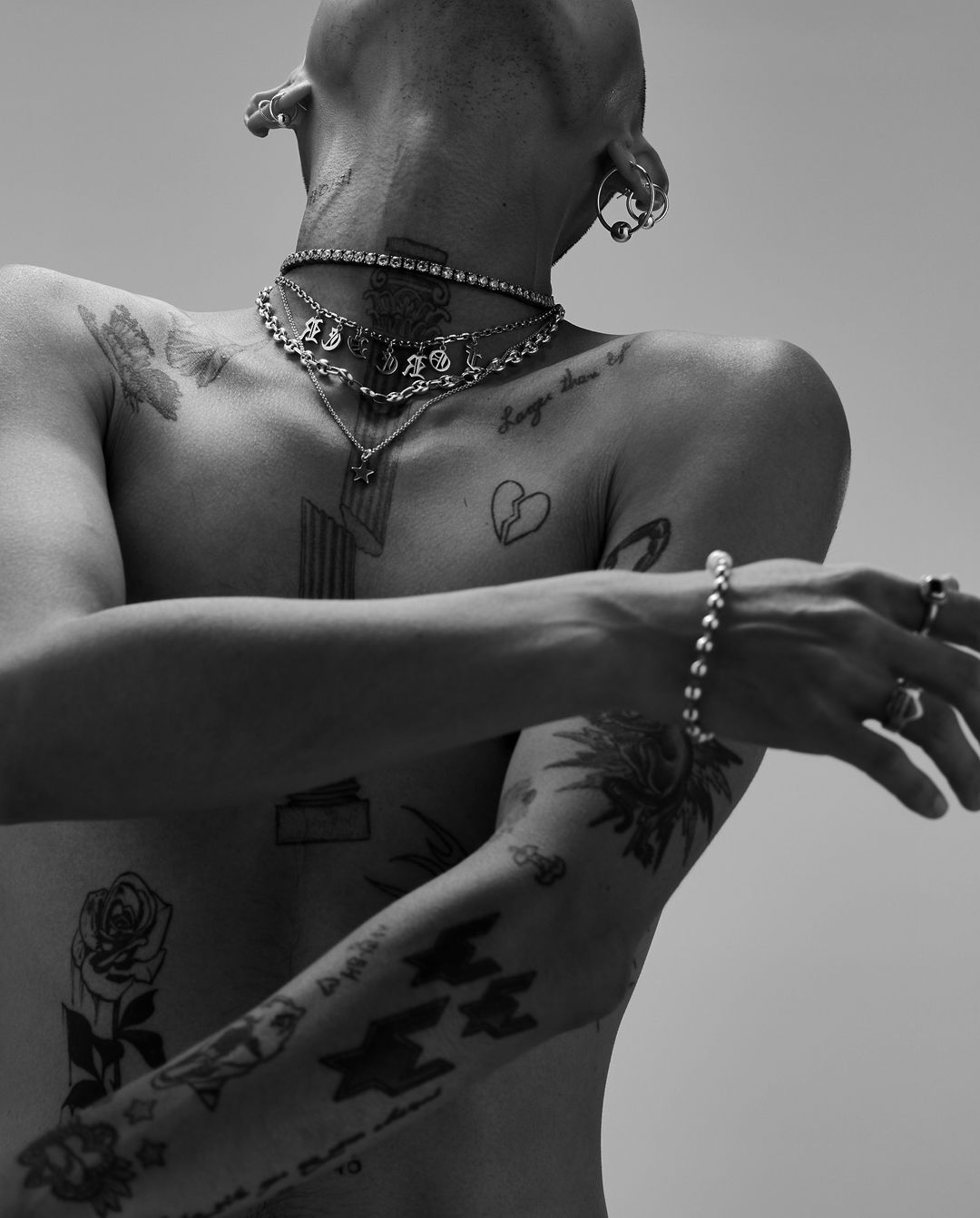 Shirtless male torso with arm tattoos, silver chains, and jewelry in a close-up black and white studio shot. Light background.