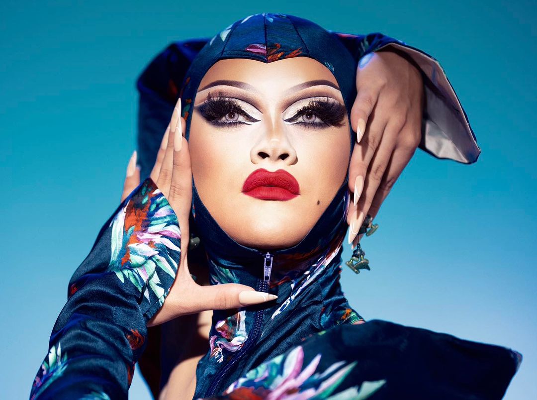 A person with dramatic makeup, red lips, and long nails poses with their hands near their face against a bright blue backdrop.