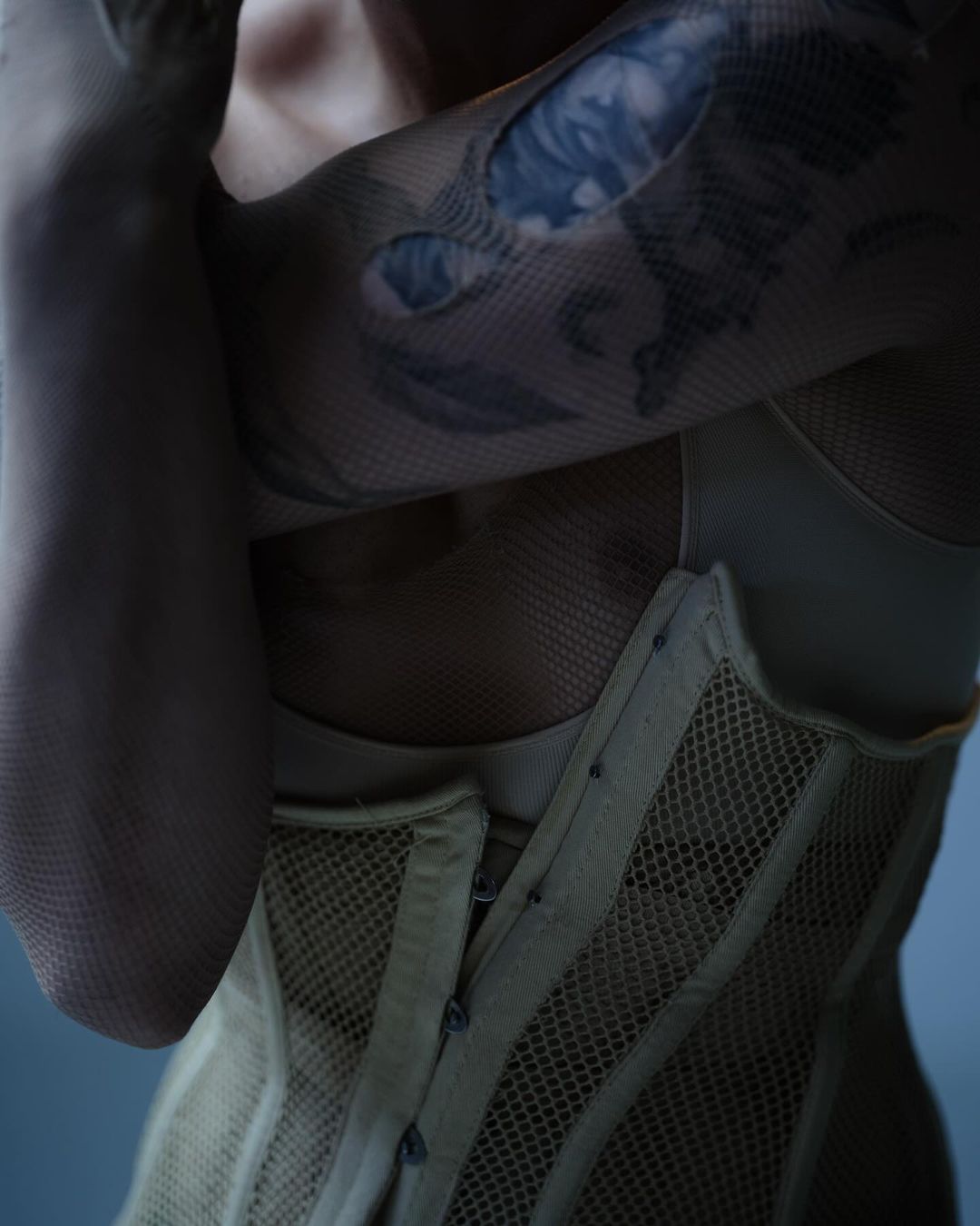 Close-up of a woman with a tattoo on her arm, wearing a beige corset and fishnet sleeves against a soft blue background.