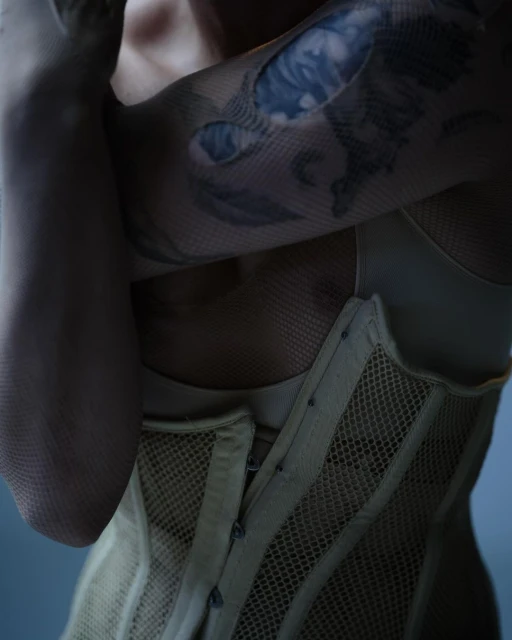 Close-up of a woman with a tattoo on her arm, wearing a beige corset and fishnet sleeves against a soft blue background.