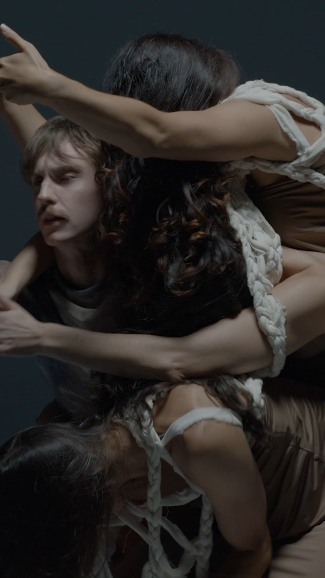 Multiple people in a close-up composition, featuring three figures intertwined and interacting with a rope element against a gray backdrop.