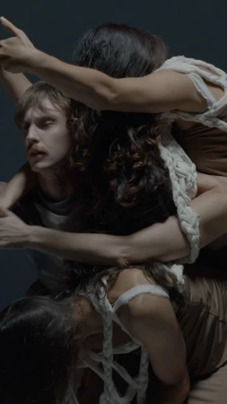 Multiple people in a close-up composition, featuring three figures intertwined and interacting with a rope element against a gray backdrop.