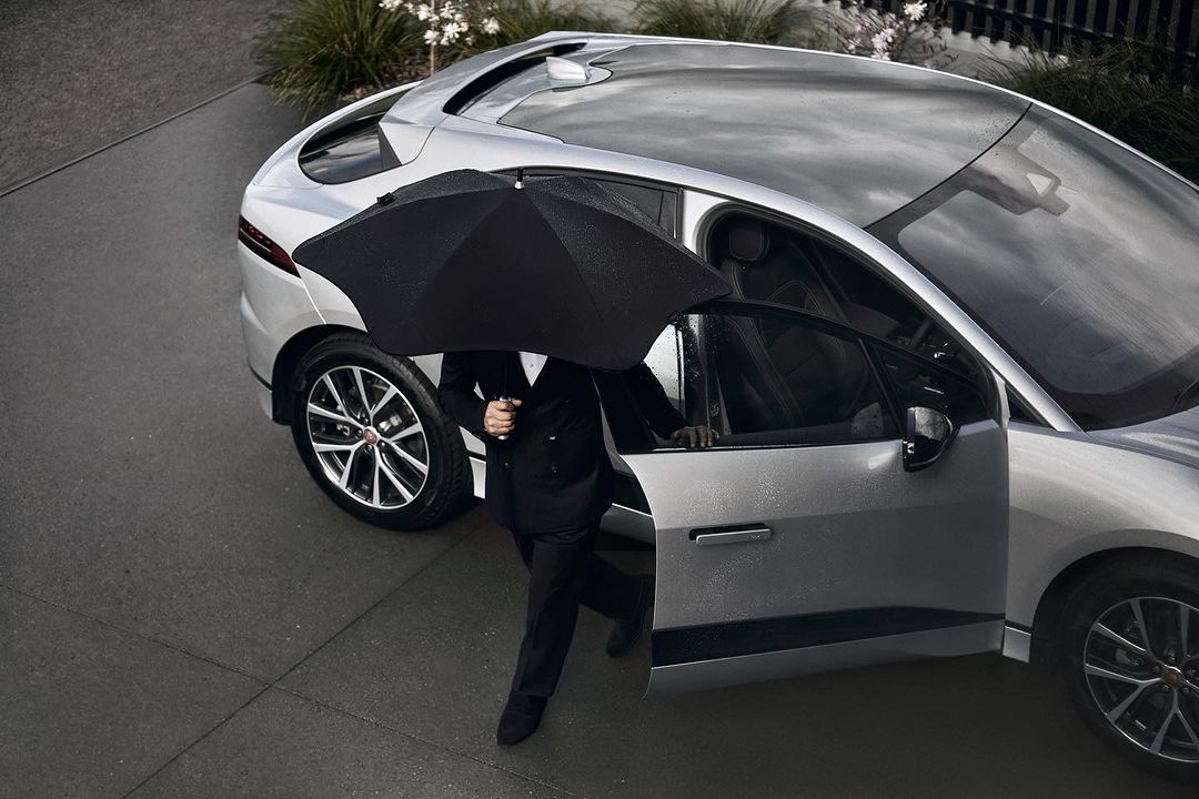 Silver SUV parked outdoors with a person in a suit exiting the vehicle while holding a black umbrella on a wet, overcast day.