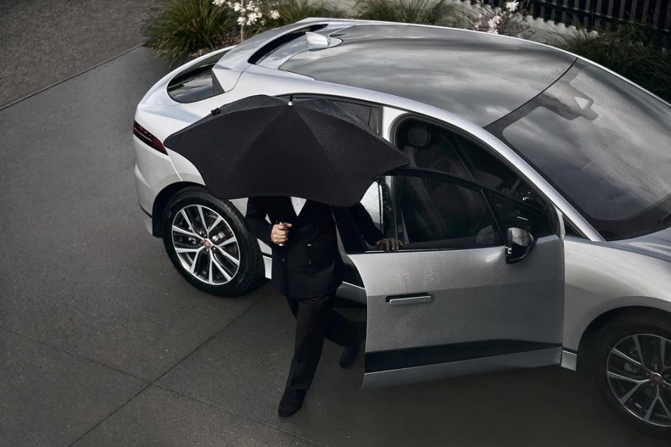 Silver SUV parked outdoors with a person in a suit exiting the vehicle while holding a black umbrella on a wet, overcast day.