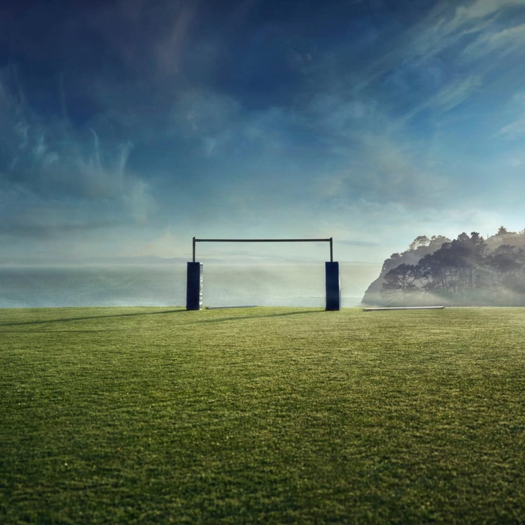 Rugby goalposts stand on a green field beneath a dramatic sky near the ocean and a tree-covered headland. The setting evokes a sense of anticipation.