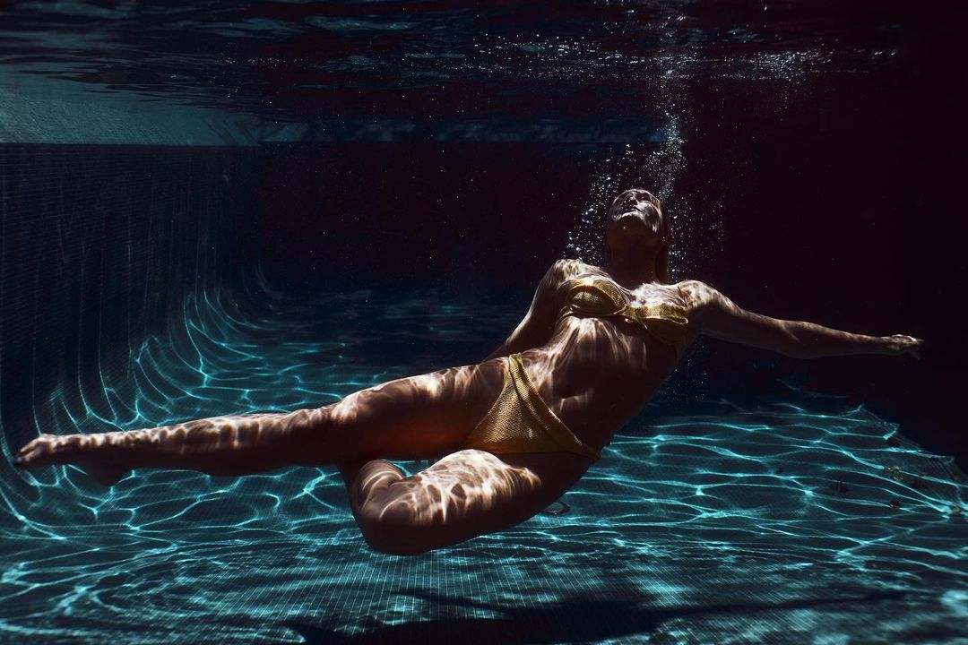 Underwater view of a woman in a yellow bikini gracefully swimming in a pool, with light patterns playing across the tiled surface.