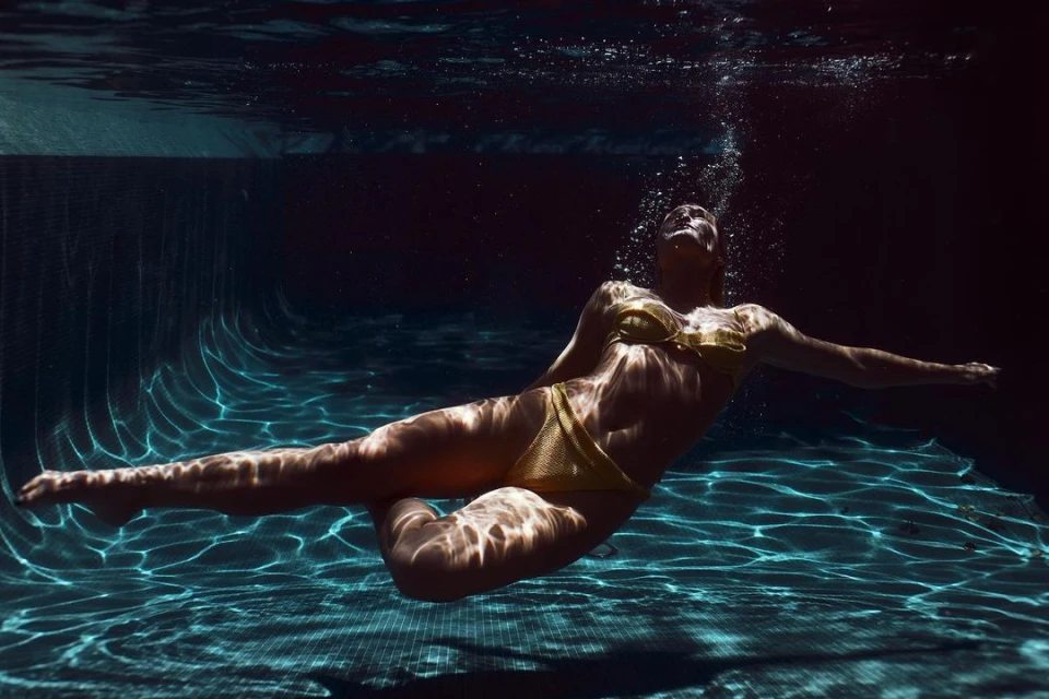 Underwater view of a woman in a yellow bikini gracefully swimming in a pool, with light patterns playing across the tiled surface.