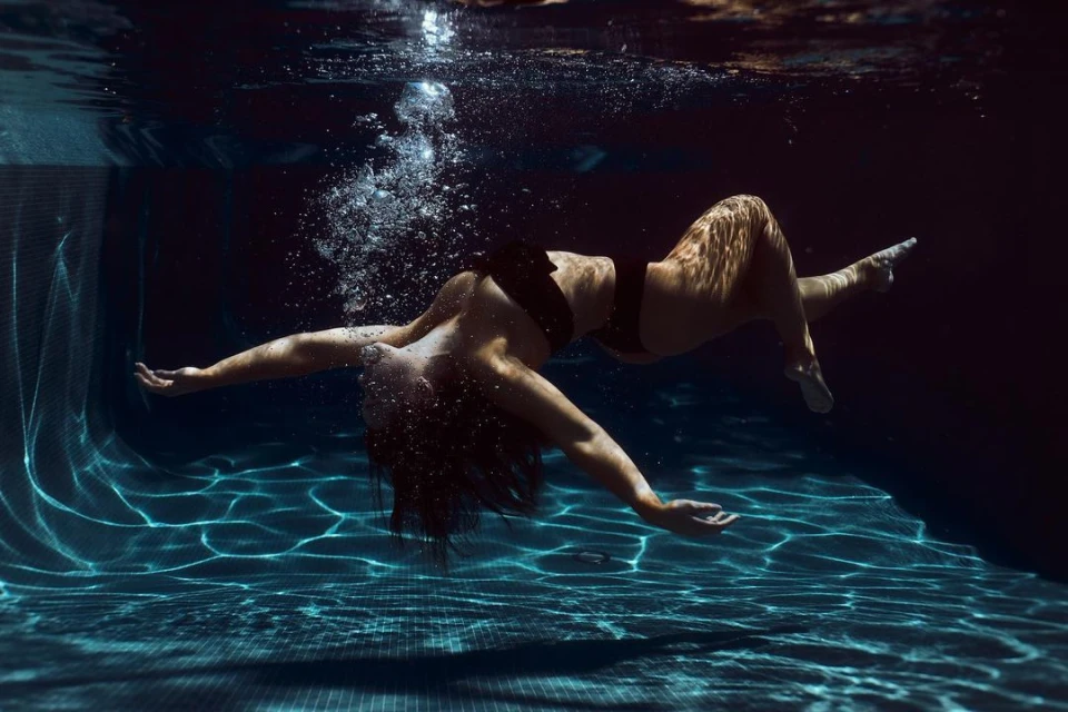 An adult female underwater with her dark hair flowing, wearing a black swimsuit, in a swimming pool with sun reflections.