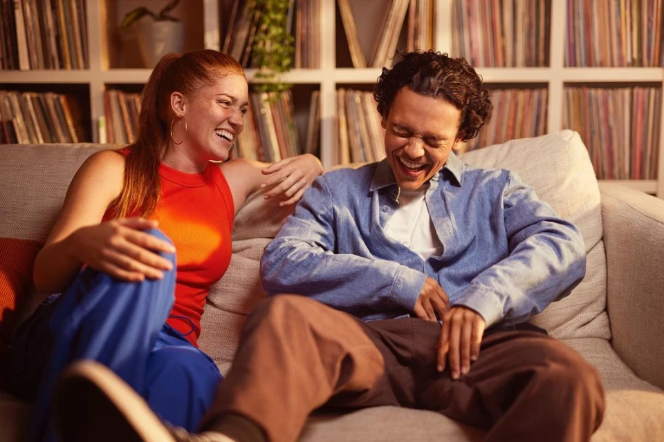 Two adults, a man and woman, laugh together on a light-colored couch in a home interior with records displayed on shelves behind them.