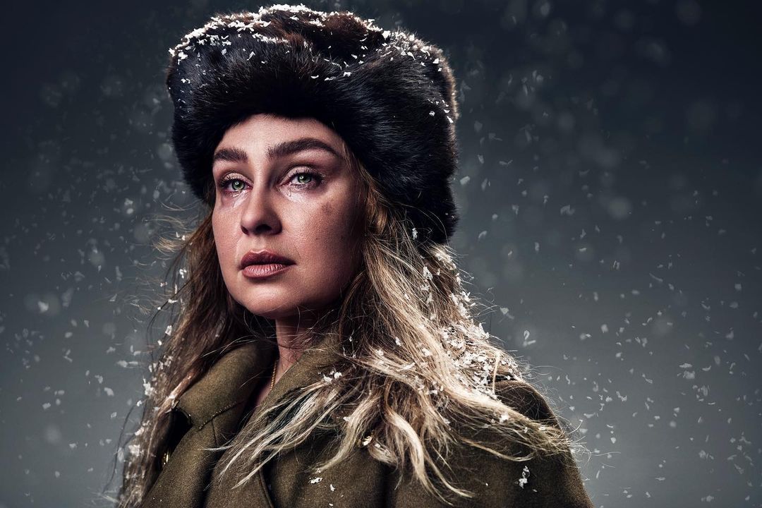 Portrait of an adult female with blonde hair wearing a fur hat and coat, looking into the distance against a grey background.