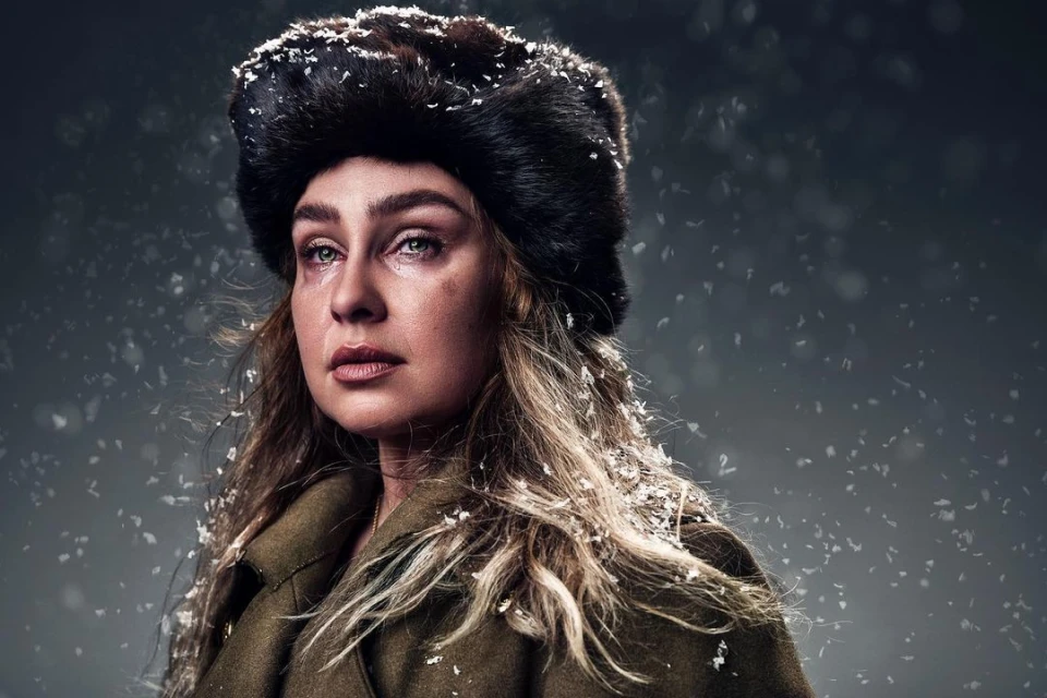 Portrait of an adult female with blonde hair wearing a fur hat and coat, looking into the distance against a grey background.