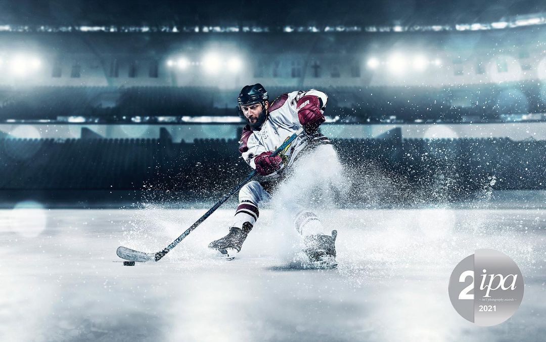 A dramatic, wide shot of a male hockey player in action on an ice rink, spraying ice with his hockey stick against a stadium backdrop.
