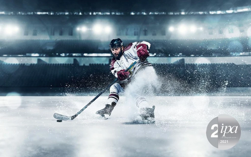 A dramatic, wide shot of a male hockey player in action on an ice rink, spraying ice with his hockey stick against a stadium backdrop.