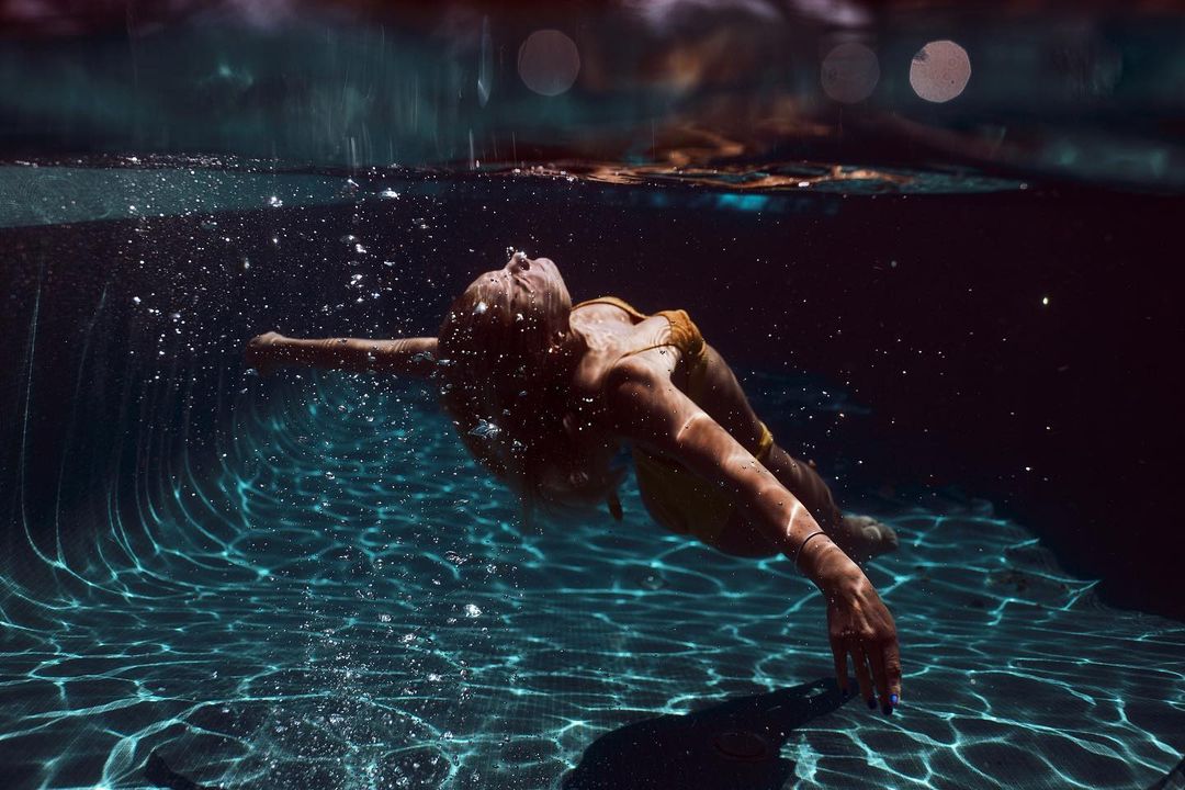 Underwater shot of an adult woman with blonde hair wearing a yellow swimsuit, swimming in a pool with rippled reflections.
