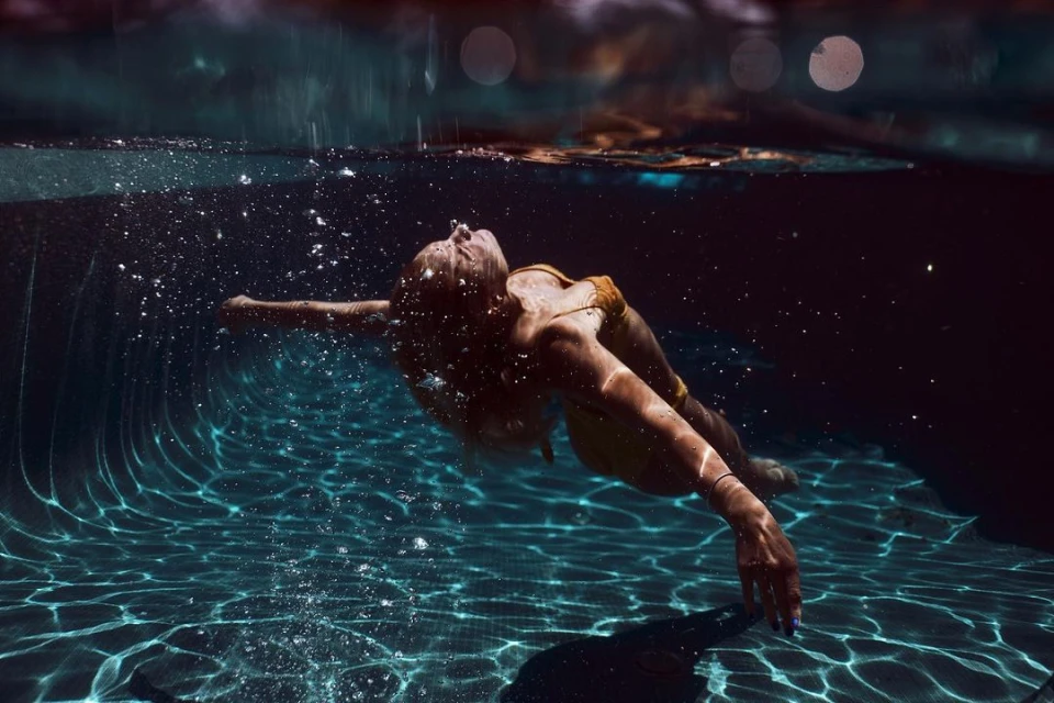 Underwater shot of an adult woman with blonde hair wearing a yellow swimsuit, swimming in a pool with rippled reflections.