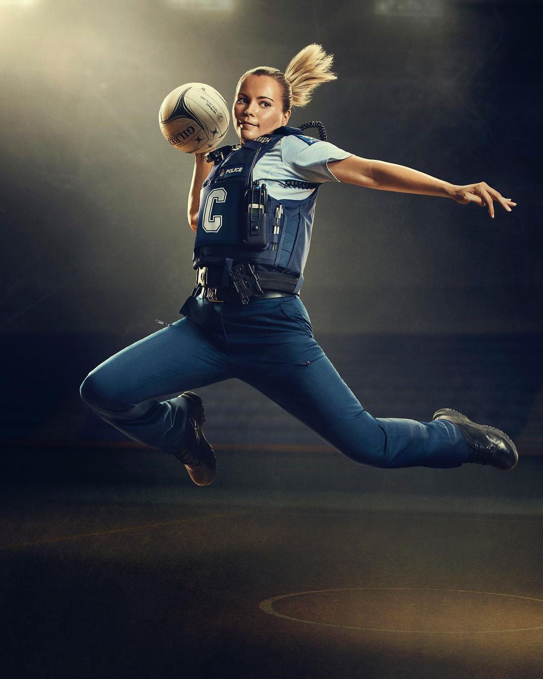 Action shot of a female adult in police tactical gear jumping in the air while throwing a netball against a dark, smoky studio backdrop.