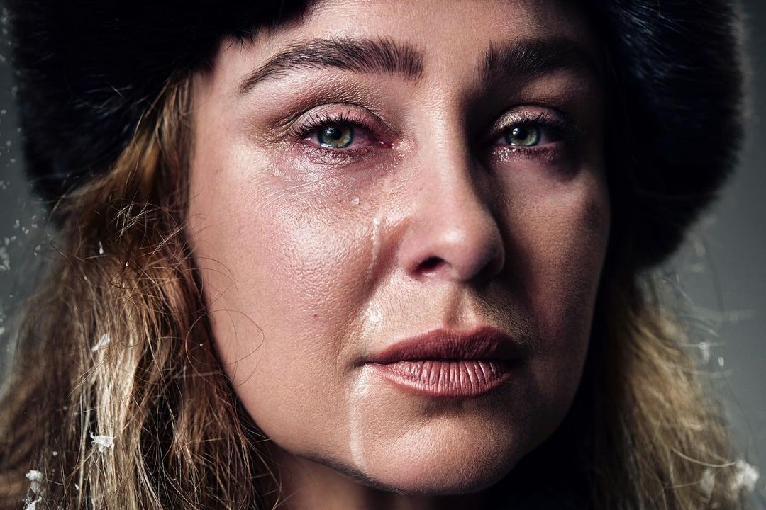 Close-up portrait of a crying adult woman with blond hair, a dark fur hat, and fair skin. Tears stream down her face against a gray background.