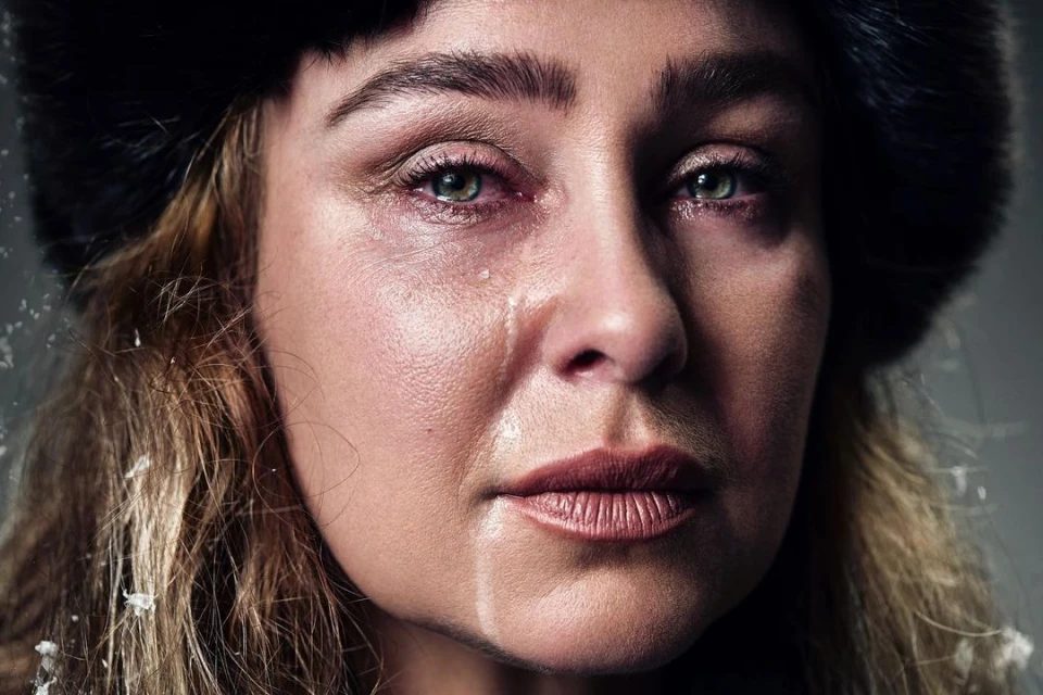 Close-up portrait of a crying adult woman with blond hair, a dark fur hat, and fair skin. Tears stream down her face against a gray background.