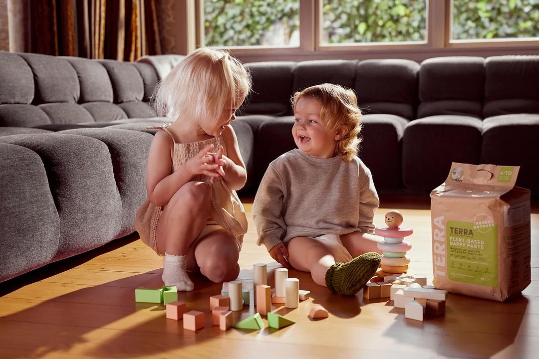 Two young children, a boy and a girl, are sitting on the floor playing with wooden blocks and toys in a brightly lit living room.