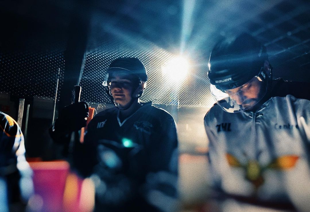 Two hockey players with helmets and jerseys stand in a dimly lit indoor ice rink with a textured wall behind them.