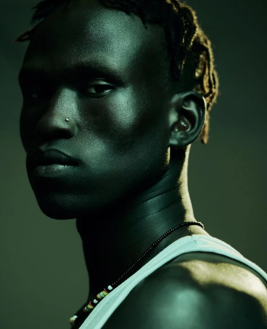 Portrait of a dark-skinned man with beaded necklaces and a nose ring, captured in a studio setting with moody, greenish lighting.