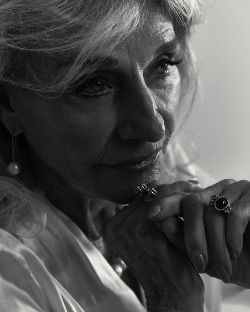 Black and white portrait of an elderly woman with blonde hair wearing jewelry, close-up shot in a brightly lit indoor setting.