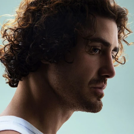 Profile portrait of a man with long curly brown hair and stubble wearing a white ribbed tank top against a light background.