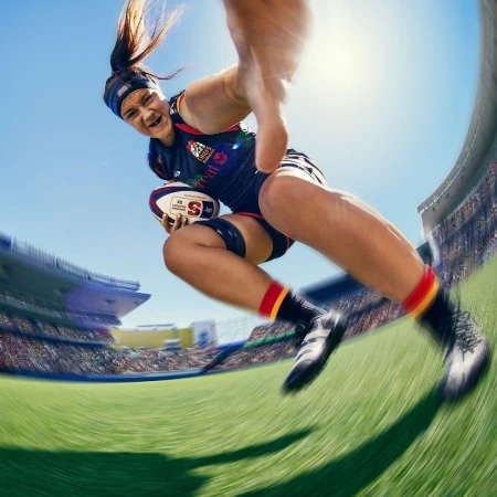 Action shot of a female rugby player in mid-air, holding the ball in a stadium setting under a clear sky, captured with a dynamic angle.