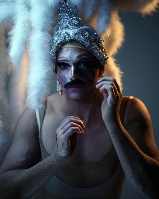 A person with a moustache wearing makeup and a jeweled headdress with feathers posing in a photo studio with dramatic lighting.