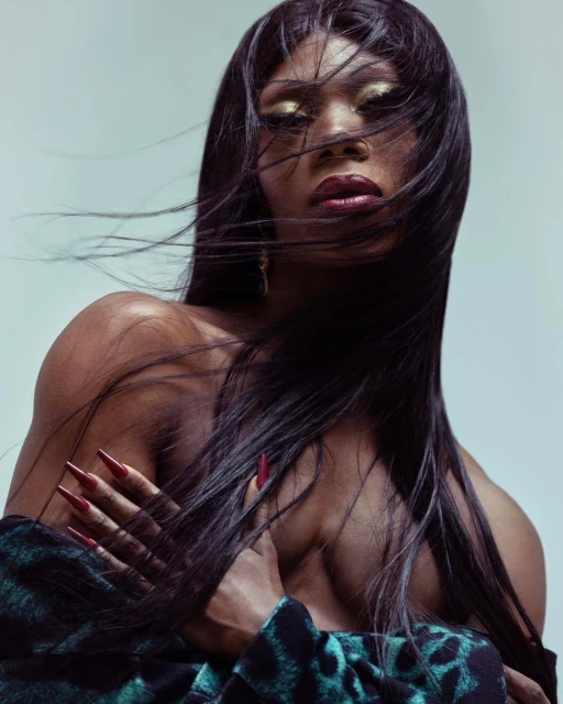 Portrait of a non-binary adult with long black hair, bold makeup, and red manicure, wearing a patterned top in a photo studio.