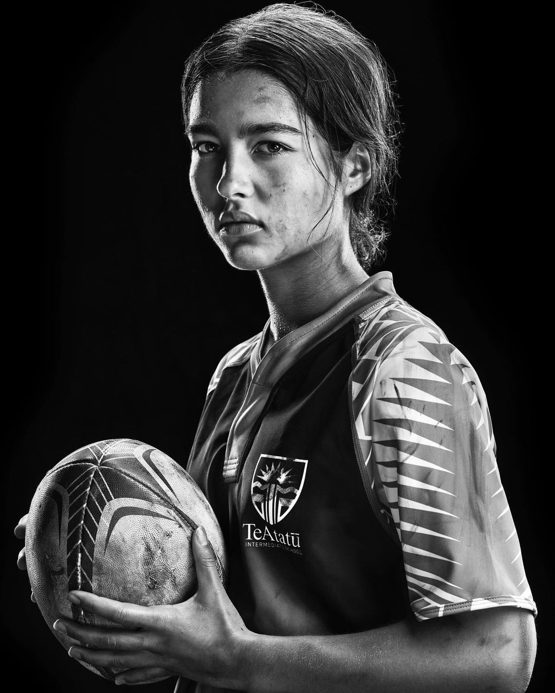 Black and white portrait of a young woman in a sports jersey holding a rugby ball, set against a dark, seamless background.