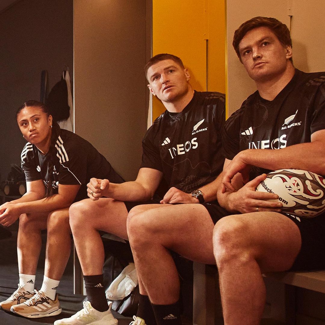 Three rugby players in team jerseys sit side-by-side in a locker room setting, holding a rugby ball and looking forward.