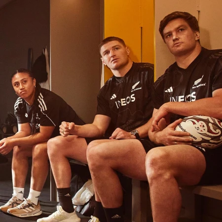 Three rugby players in team jerseys sit side-by-side in a locker room setting, holding a rugby ball and looking forward.