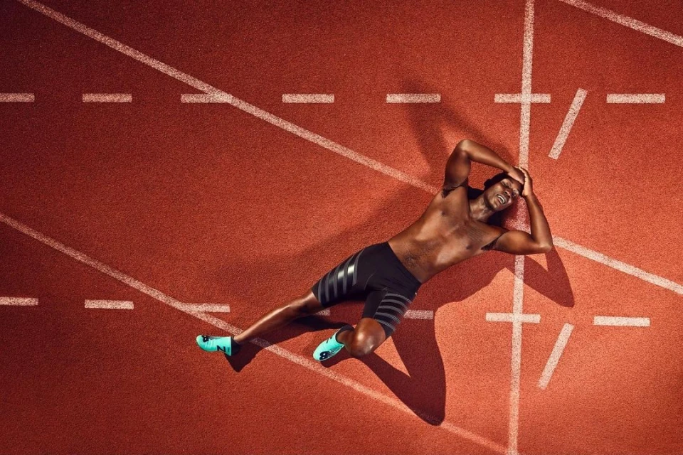 Athlete lies exhausted on a red running track with white lines, hands on his head, wearing running shoes and shorts. Top-down, eye-level view.
