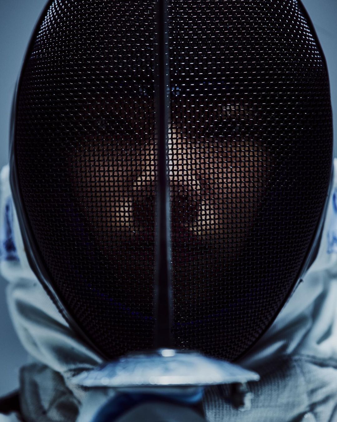 Close-up of a fencer wearing a black fencing mask and white uniform, looking directly at the camera in a studio setting.