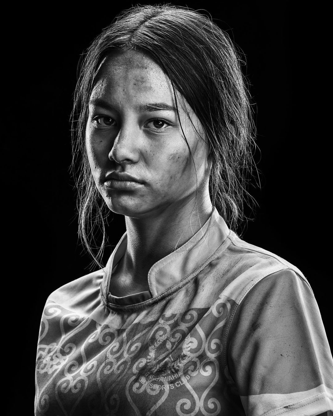 Monochrome close-up of a young adult female athlete with a serious expression, showing the details of dirt on her face.