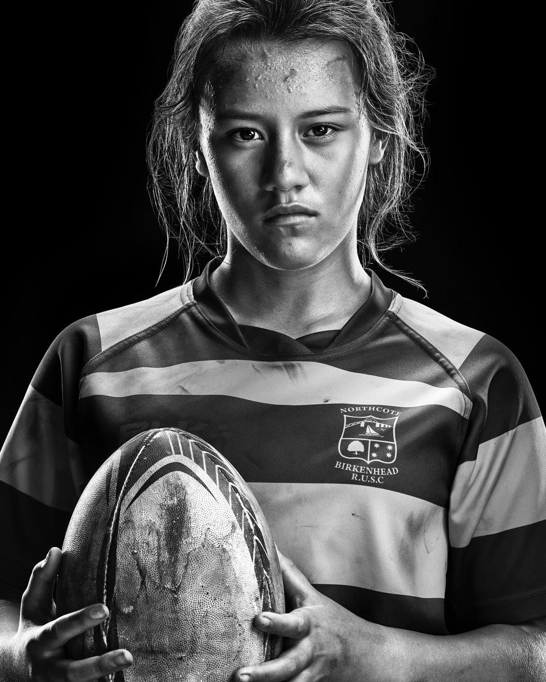 Black and white portrait of a determined young woman in a striped rugby jersey holding a worn rugby ball against a black backdrop.