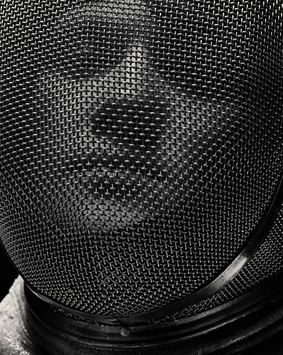 Close-up of a person wearing a fencing mask with a metallic net, with the dark background adding contrast to the metallic tones.