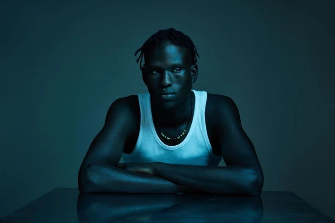 Portrait of a Black man in a white tank top with arms crossed, photographed in cool blue studio lighting by Sacha Stejko