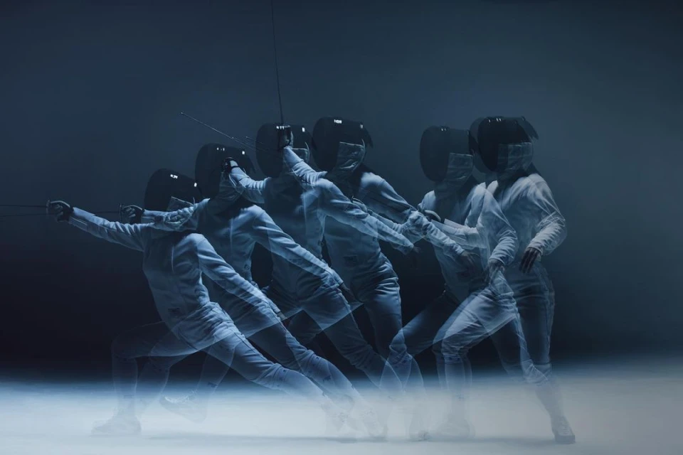 Multiple exposures of a fencer in white fencing gear and black mask performing a fencing move against a dark background.