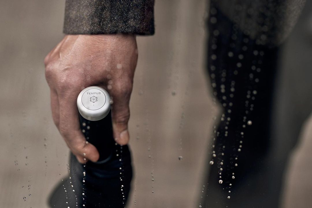 Close-up of a hand holding a black bottle with a white cap, showcasing the brand logo and subtle water droplets on a clear surface.