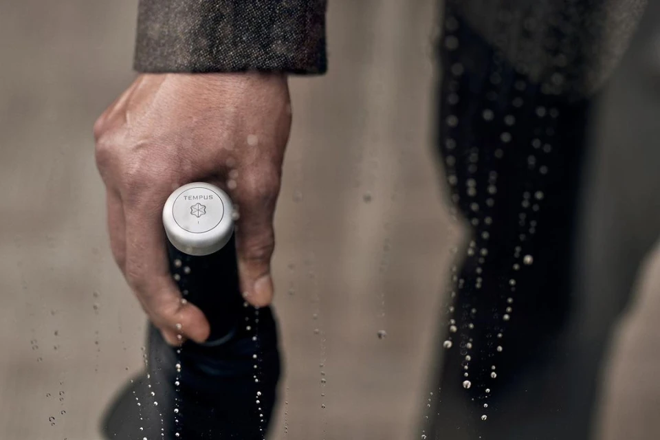 Close-up of a hand holding a black bottle with a white cap, showcasing the brand logo and subtle water droplets on a clear surface.