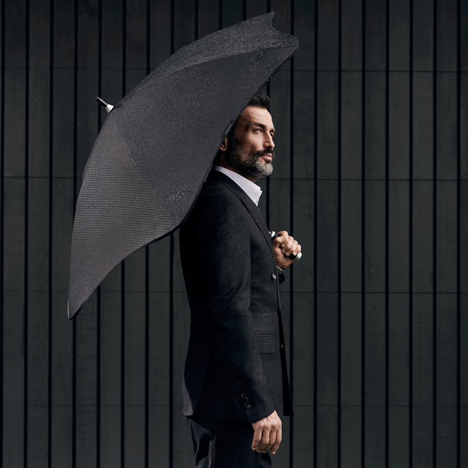 Elegant adult man in a tailored suit stands outdoors with a dark umbrella against a striped background, exuding a sophisticated style.