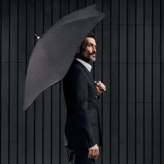 Elegant adult man in a tailored suit stands outdoors with a dark umbrella against a striped background, exuding a sophisticated style.