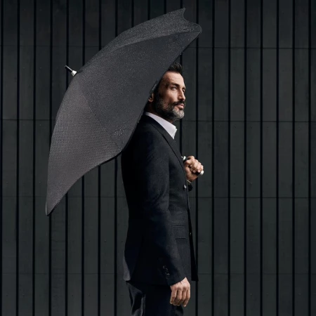 Elegant adult man in a tailored suit stands outdoors with a dark umbrella against a striped background, exuding a sophisticated style.