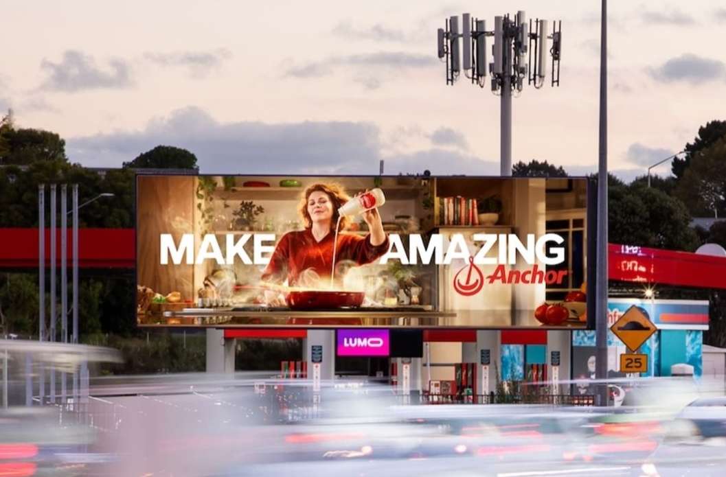 Billboard displaying woman pouring liquid into pan in kitchen scene, promoting Anchor brand, against urban background with cars and cell tower.