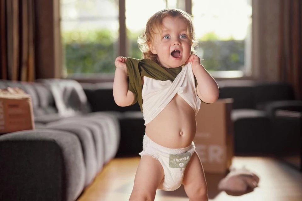 Toddler with blonde hair enthusiastically removing a green t-shirt indoors, wearing a diaper in a brightly lit living room scene.