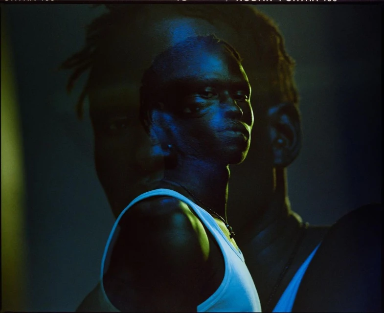 Portrait of a Black adult male in a white tank top, with blue and yellow lighting, and another human silhouetted in the background.