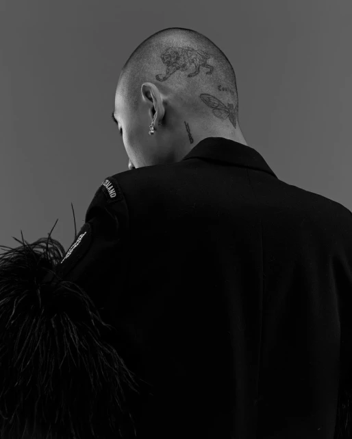 Black and white portrait of a bald adult male in a dark jacket with feathered accents, showcasing tattoos on his head and neck.